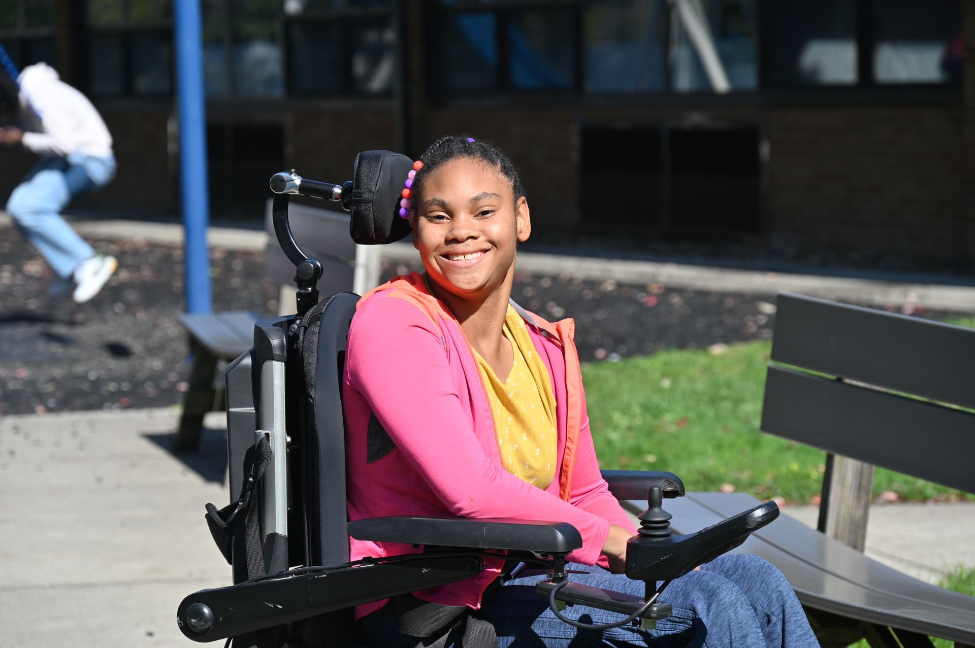 Smiling person in a wheelchair at a park, wearing a pink jacket and yellow shirt; person swinging in the background.