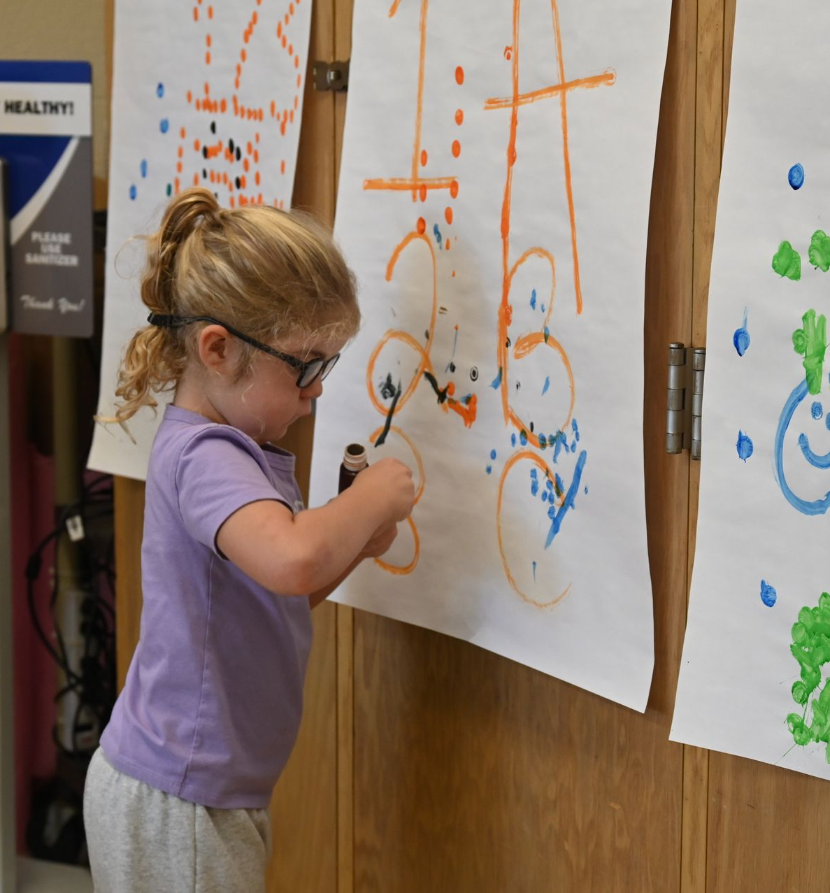 A child with glasses draws on paper attached to a wooden cabinet using a black marker.