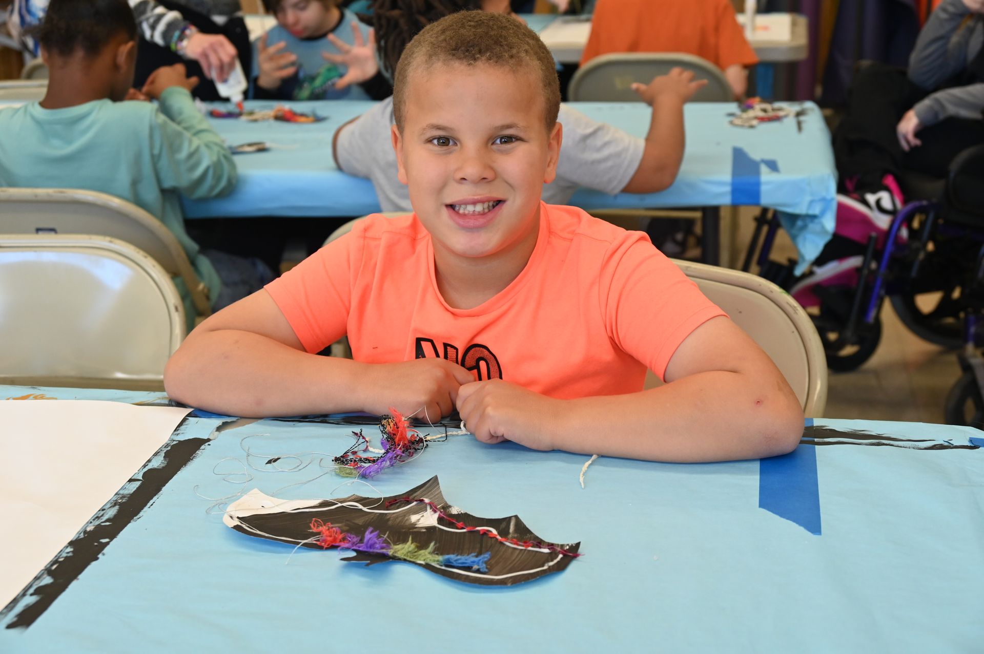 Boy smiles at the camera, art project on table. Blue tablecloth. Other children in the background.