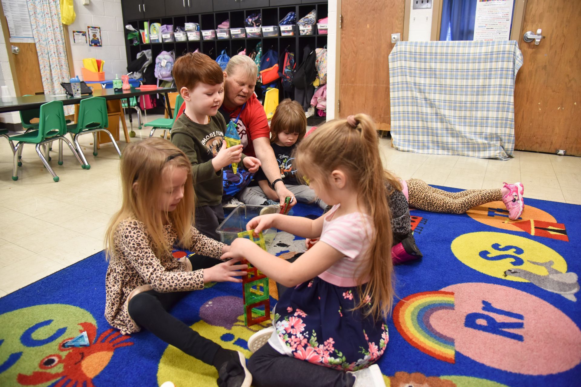 Children and teacher build with blocks on a colorful rug in a classroom.
