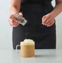 Person sprinkles dark powder onto whipped cream on coffee in a glass mug.