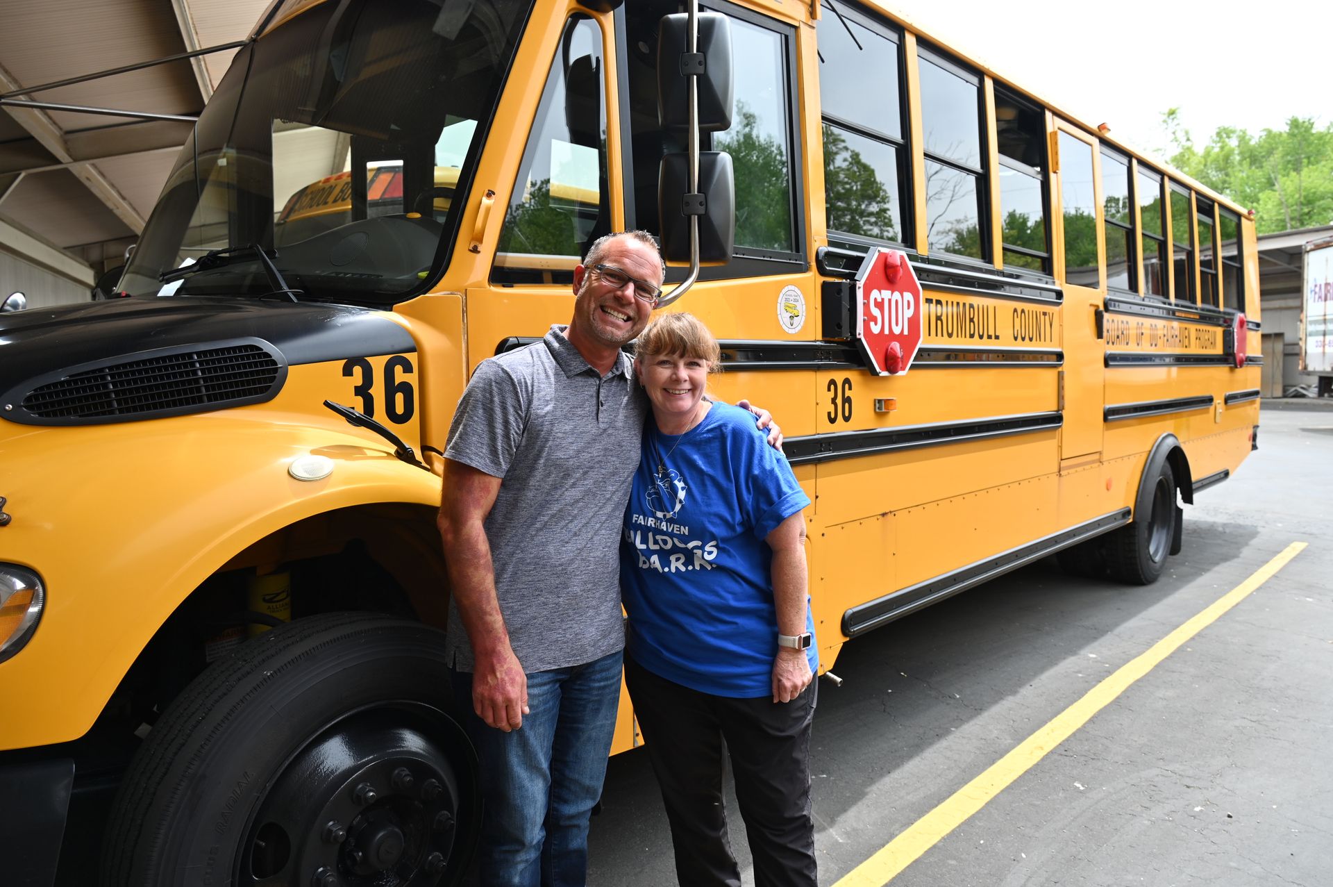 Two people pose in front of a yellow school bus with 