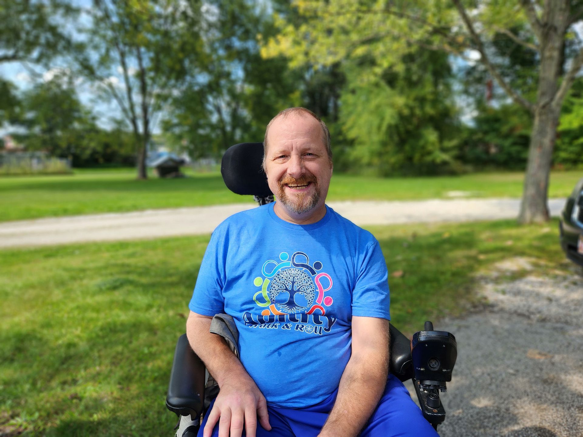 Man in a wheelchair smiles outdoors, wearing a blue t-shirt with a colorful design. Sunny, green background.