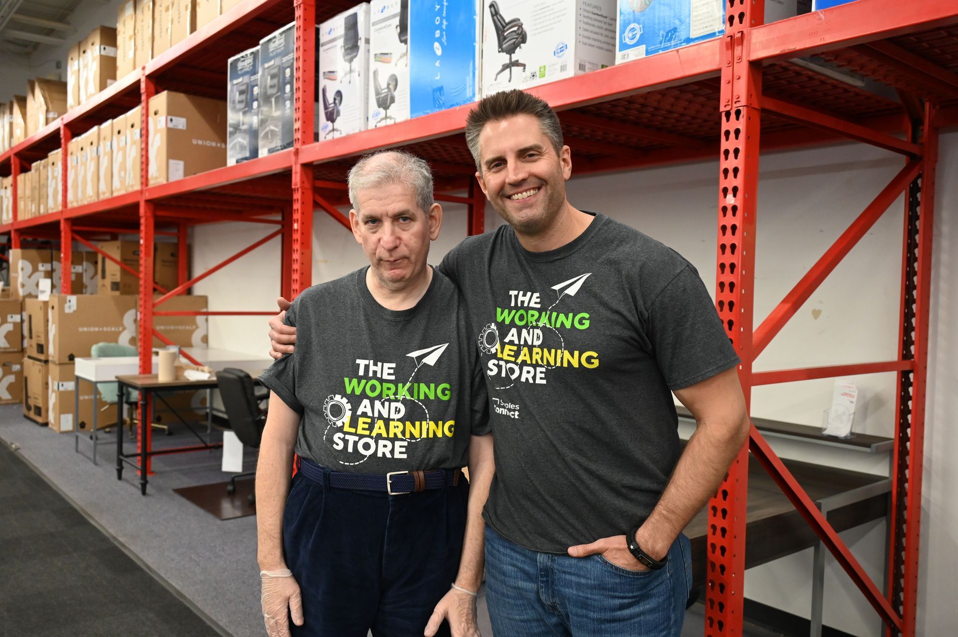 Two men in matching shirts with arms around each other in a warehouse setting, smiling.