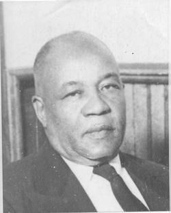 Portrait of a man in a dark suit and tie, seated indoors against wood paneling, with a neutral expression