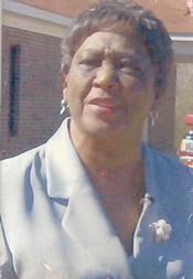 Woman in a light blue suit outdoors, wearing a white corsage and earrings, with a brick building behind her