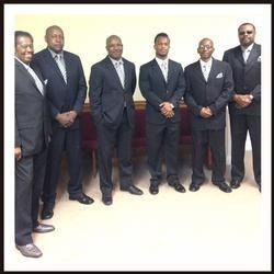 Six men in dark suits stand together indoors, posing formally against a light wall.