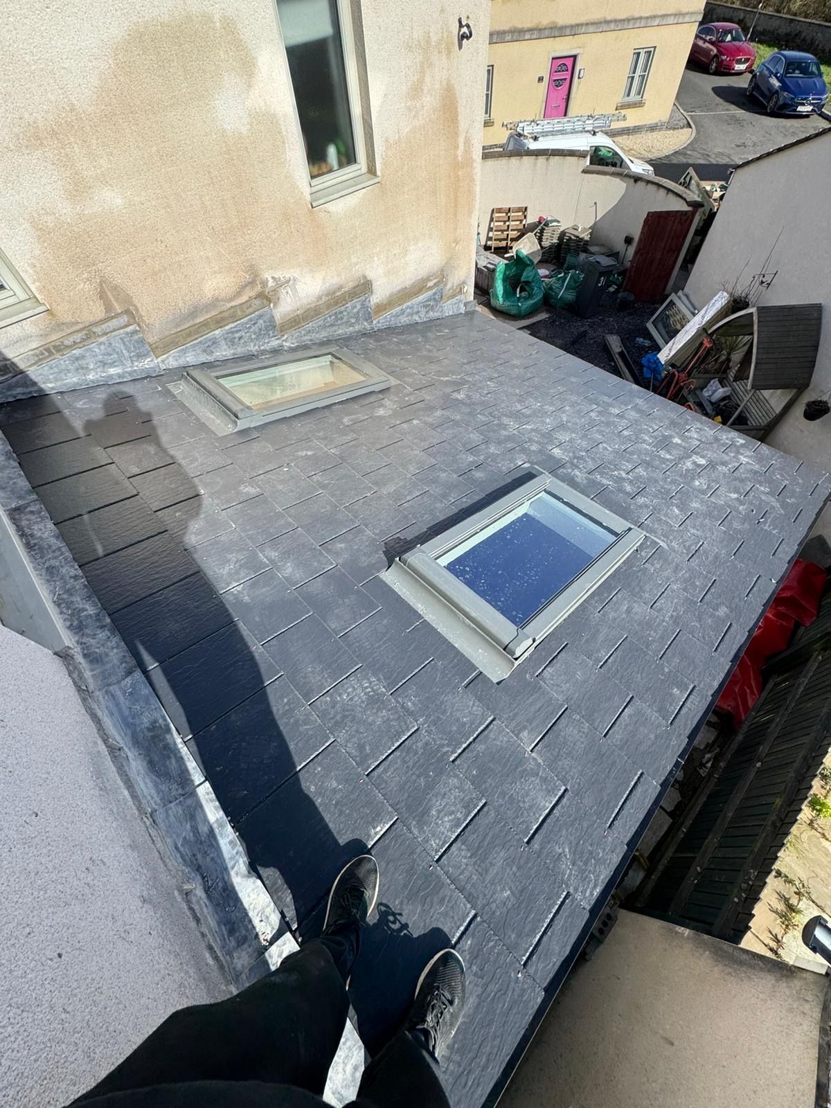 Person on a gray shingled roof with two skylights beside a beige building and parked cars below