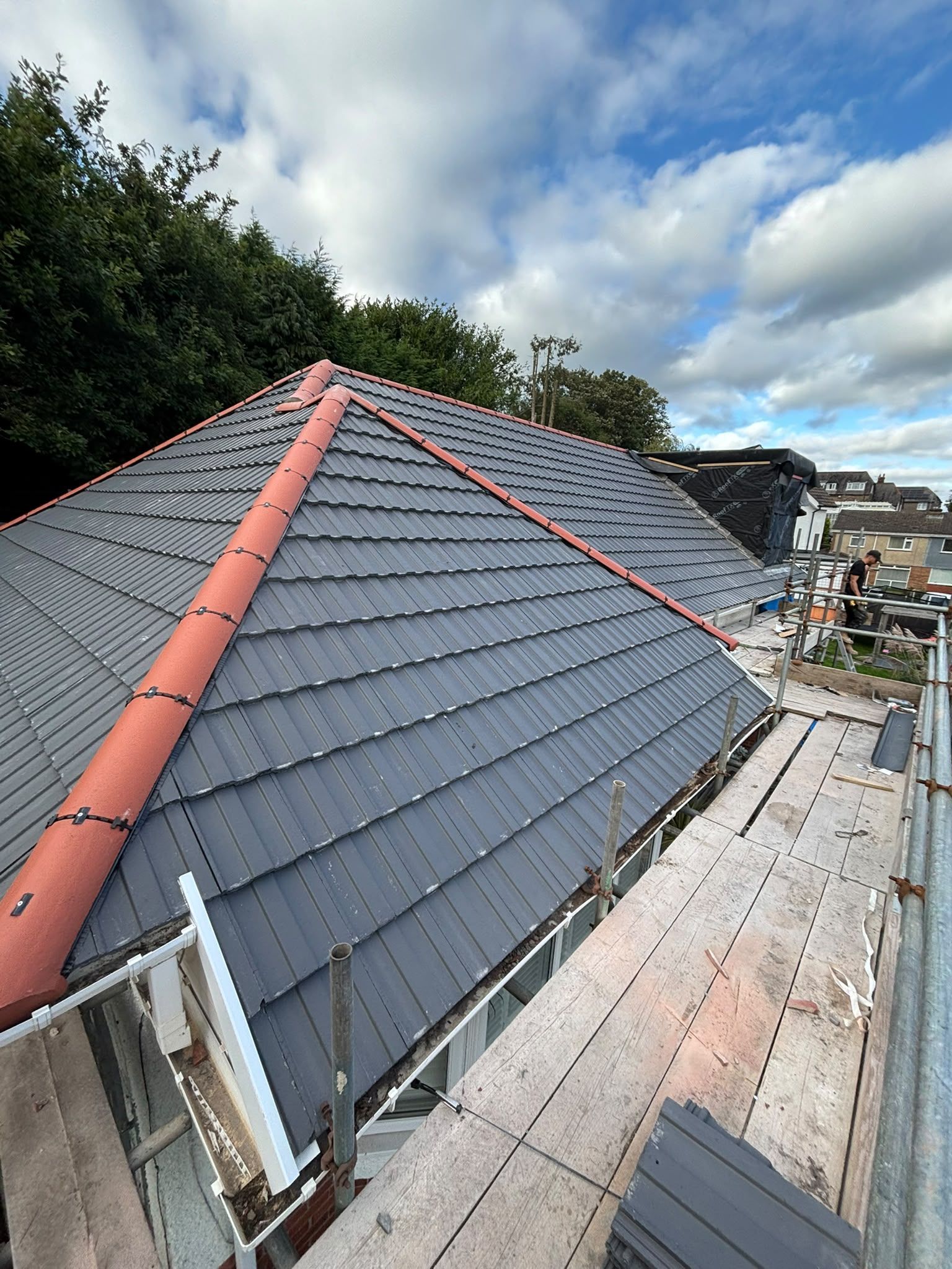 Roof under construction with dark gray tiles, red ridge trim, and scaffolding beside a hillside homesite
