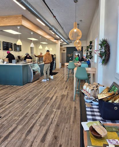 Bright café interior with teal chairs, wood floors, hanging lights, and a counter with a few customers.
