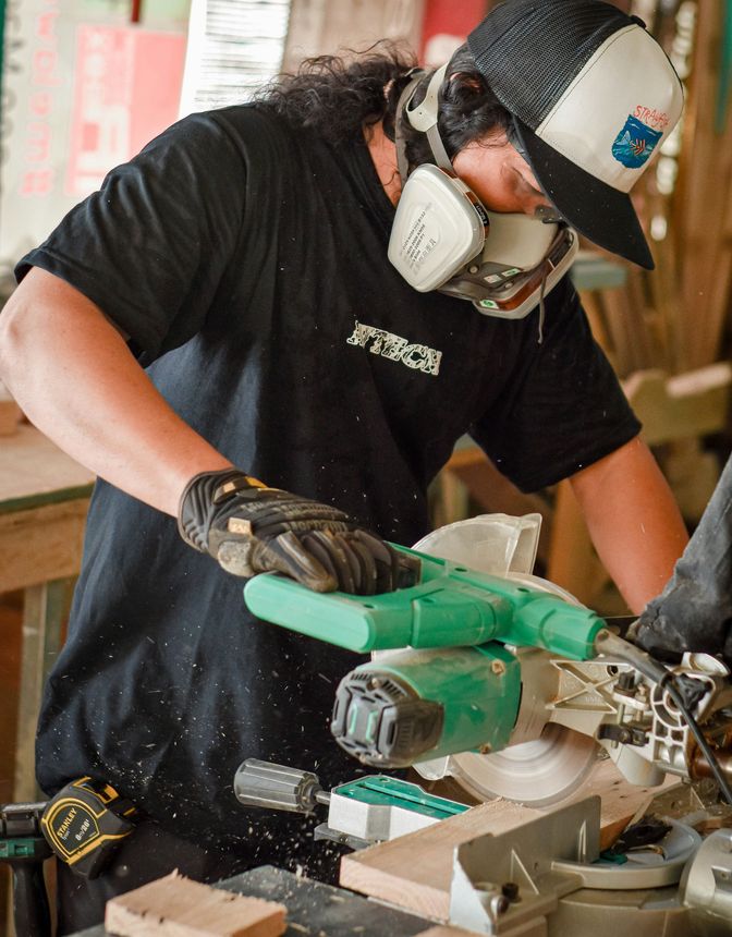 Worker using a green power saw to cut wood in a workshop, wearing gloves, mask, and hard hat