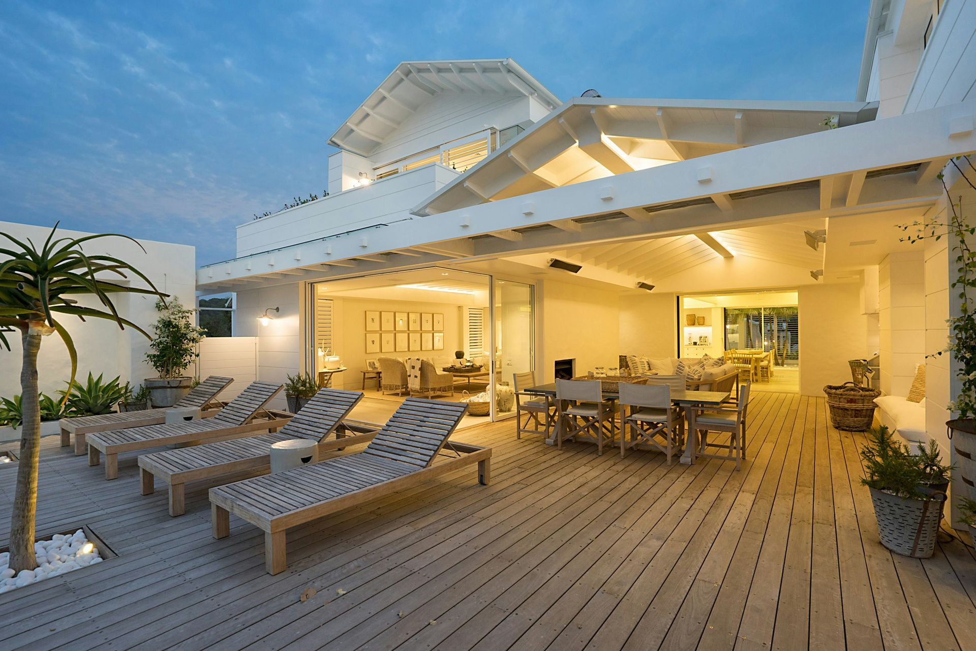 Sunlit beachfront deck with lounge chairs, dining tables, and a white wooden pavilion at dusk