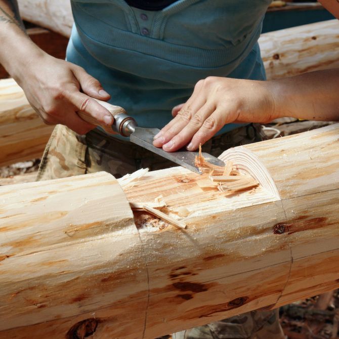 Hands carving a wooden log with a chisel and tool, creating wood shavings.