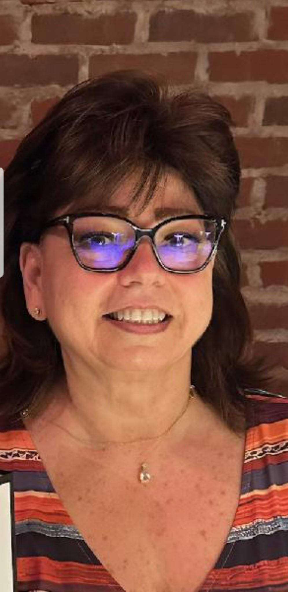 Portrait of a smiling woman with glasses in front of a brick wall, wearing a striped top.