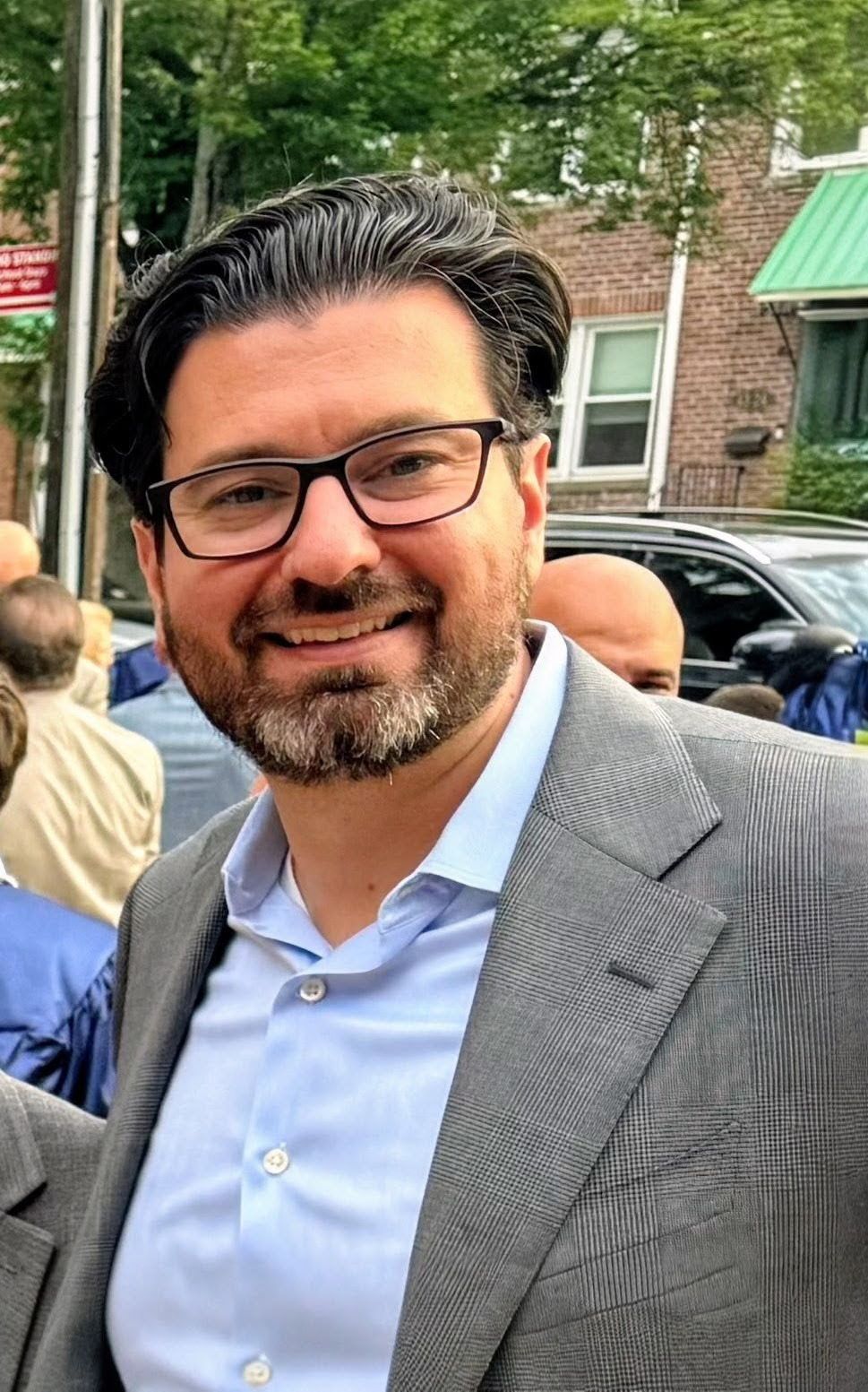 Smiling man in glasses and gray blazer outdoors on a tree-lined street