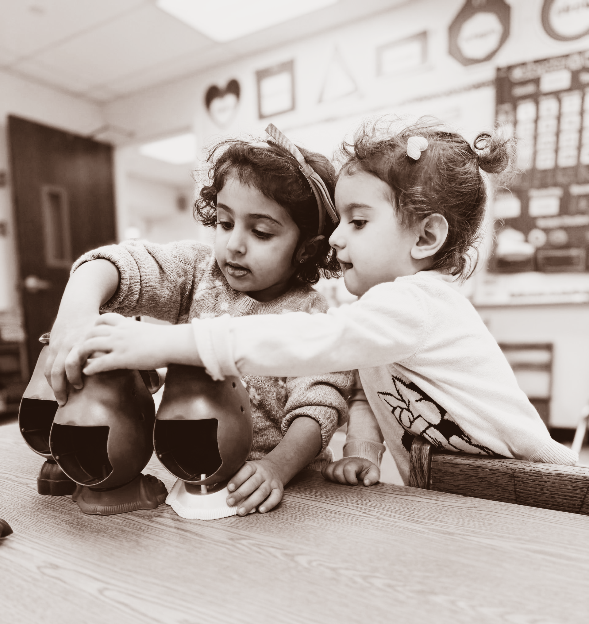 Two children sitting at a table, one helping the other with a shoe in a classroom-like room.