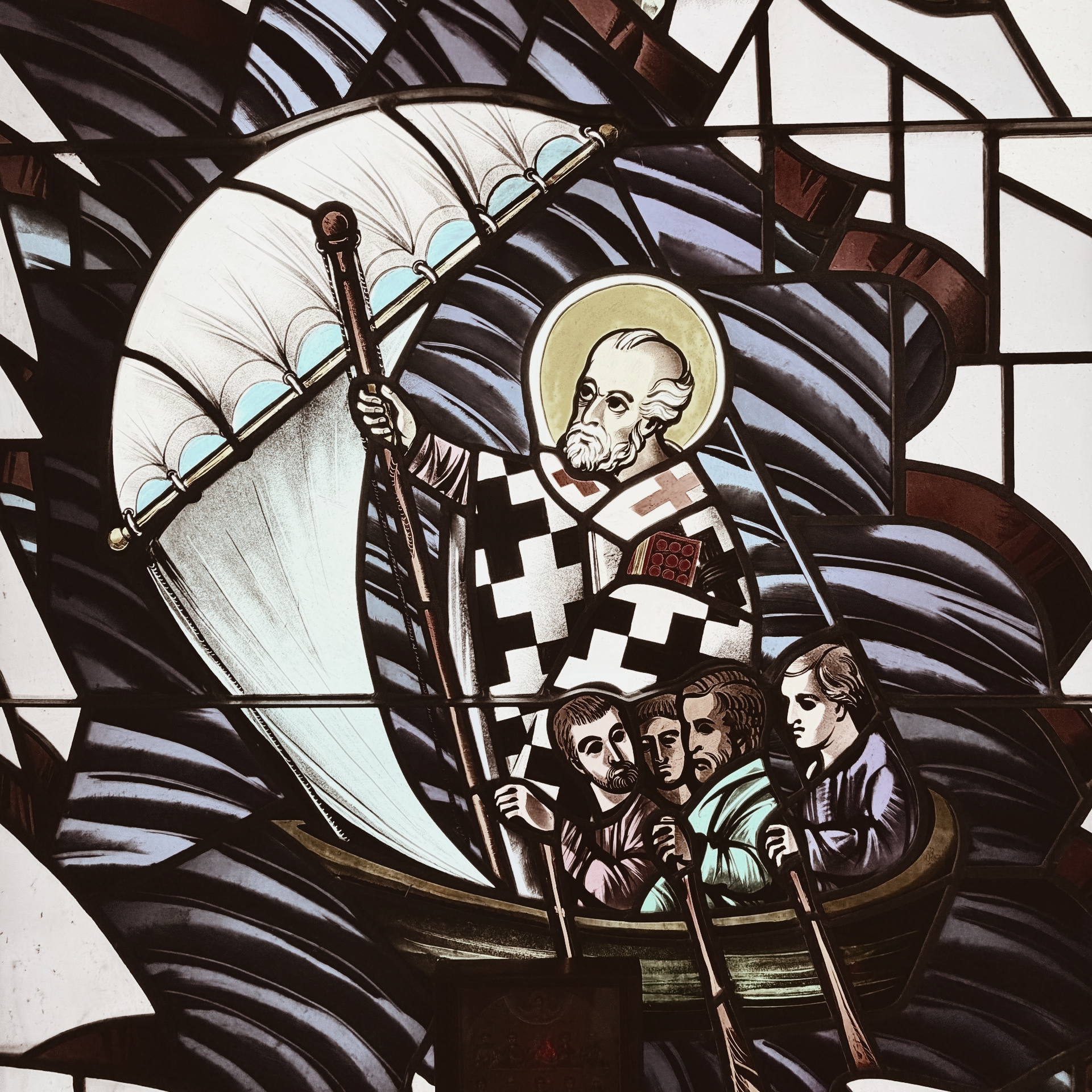 Stained glass of a robed saint with halo, raising a staff above kneeling figures in a boat-like scene