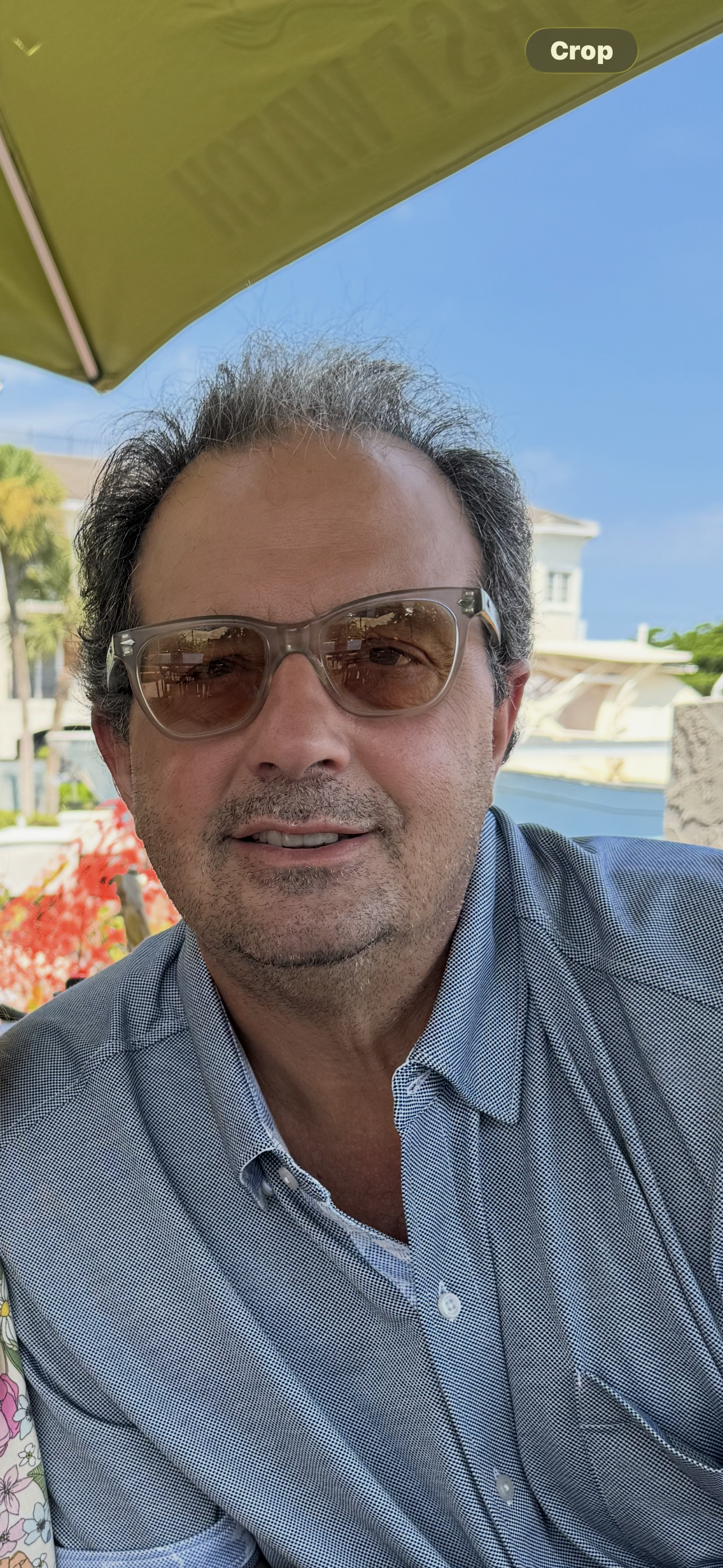 Man wearing sunglasses and a checkered shirt outdoors under a green umbrella, smiling at the camera
