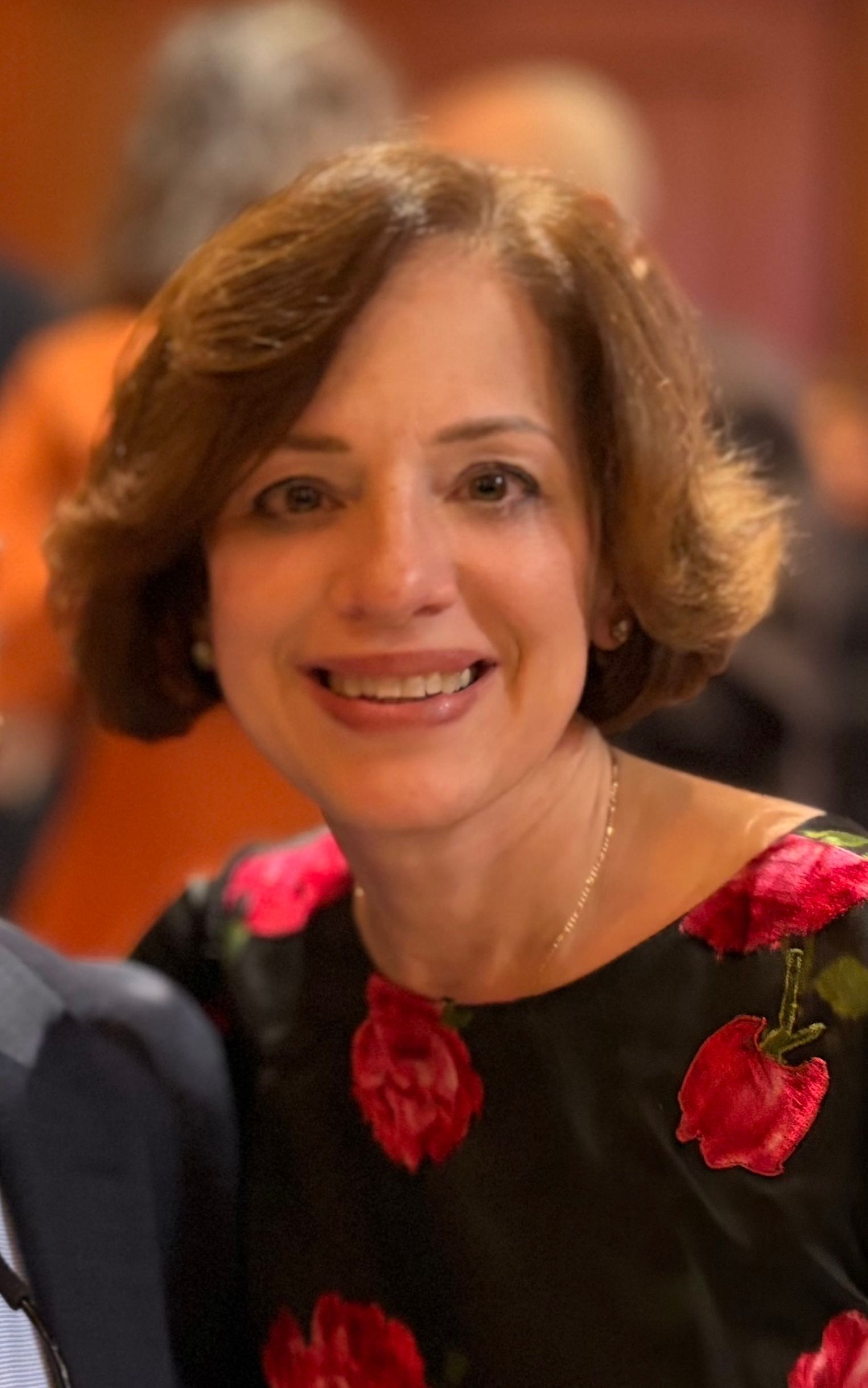 Smiling woman in a black dress with red floral accents at an indoor event, against a warm orange background