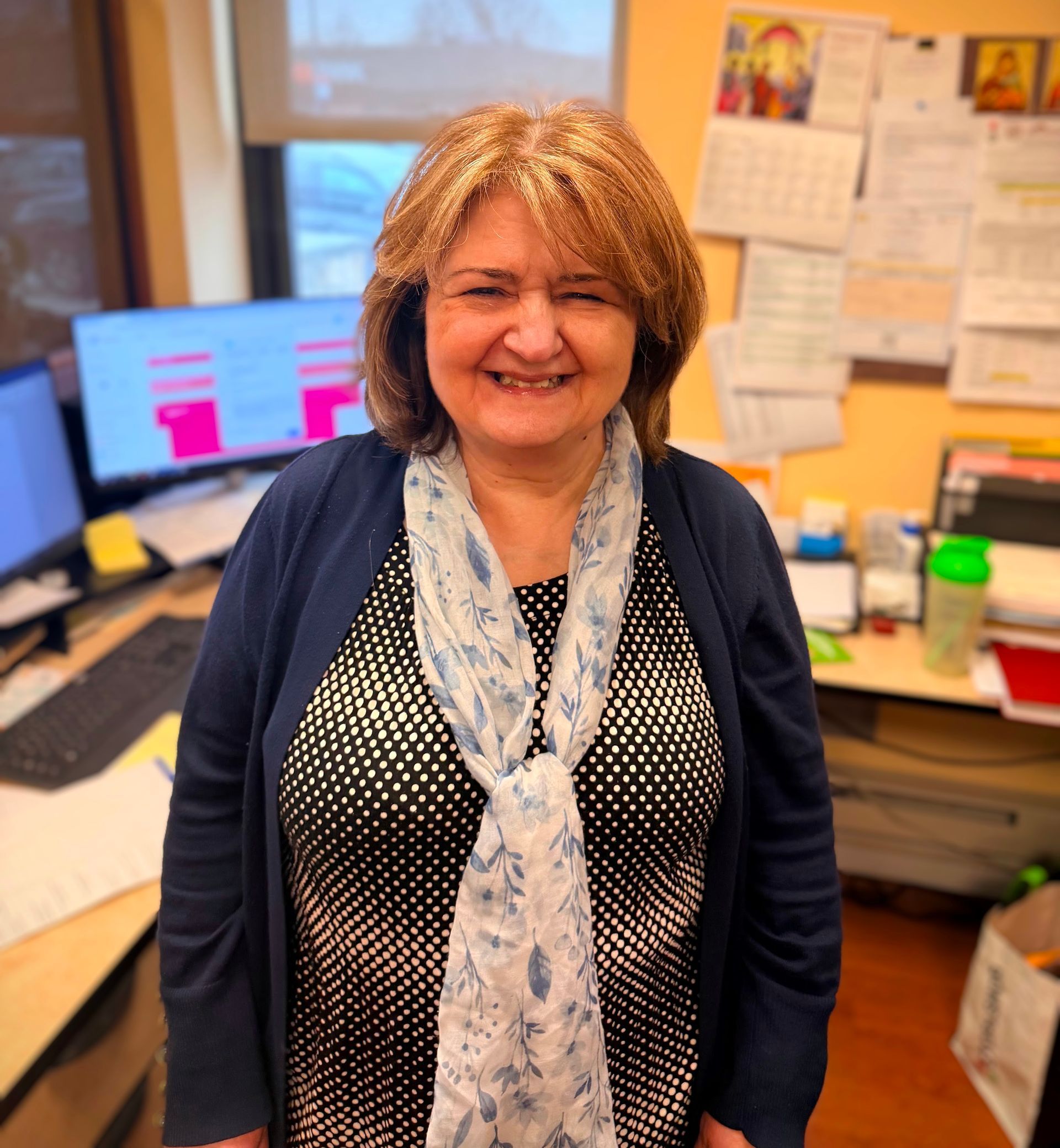 Smiling woman standing in an office, wearing a dark cardigan and patterned scarf, with desks and monitors behind her