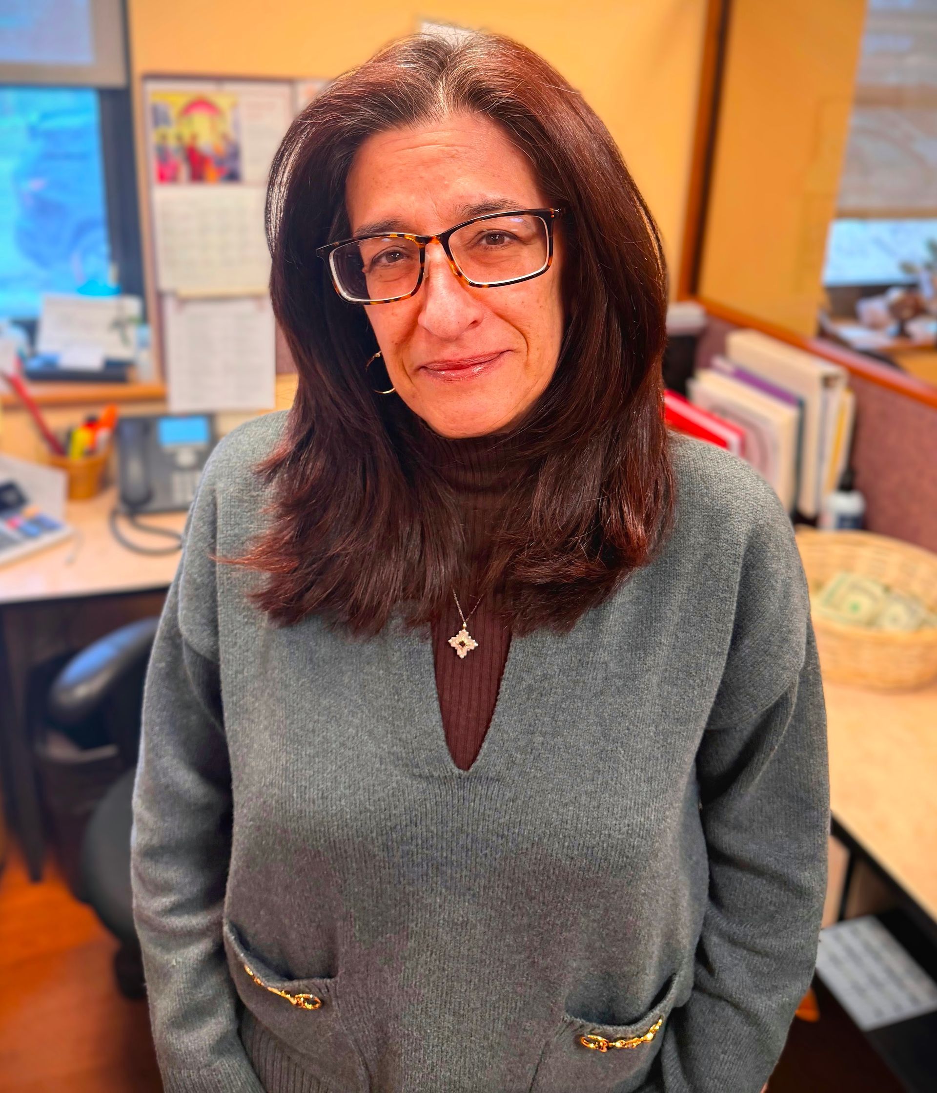 Woman smiling in an office, wearing glasses and a gray sweater with gold zippers.