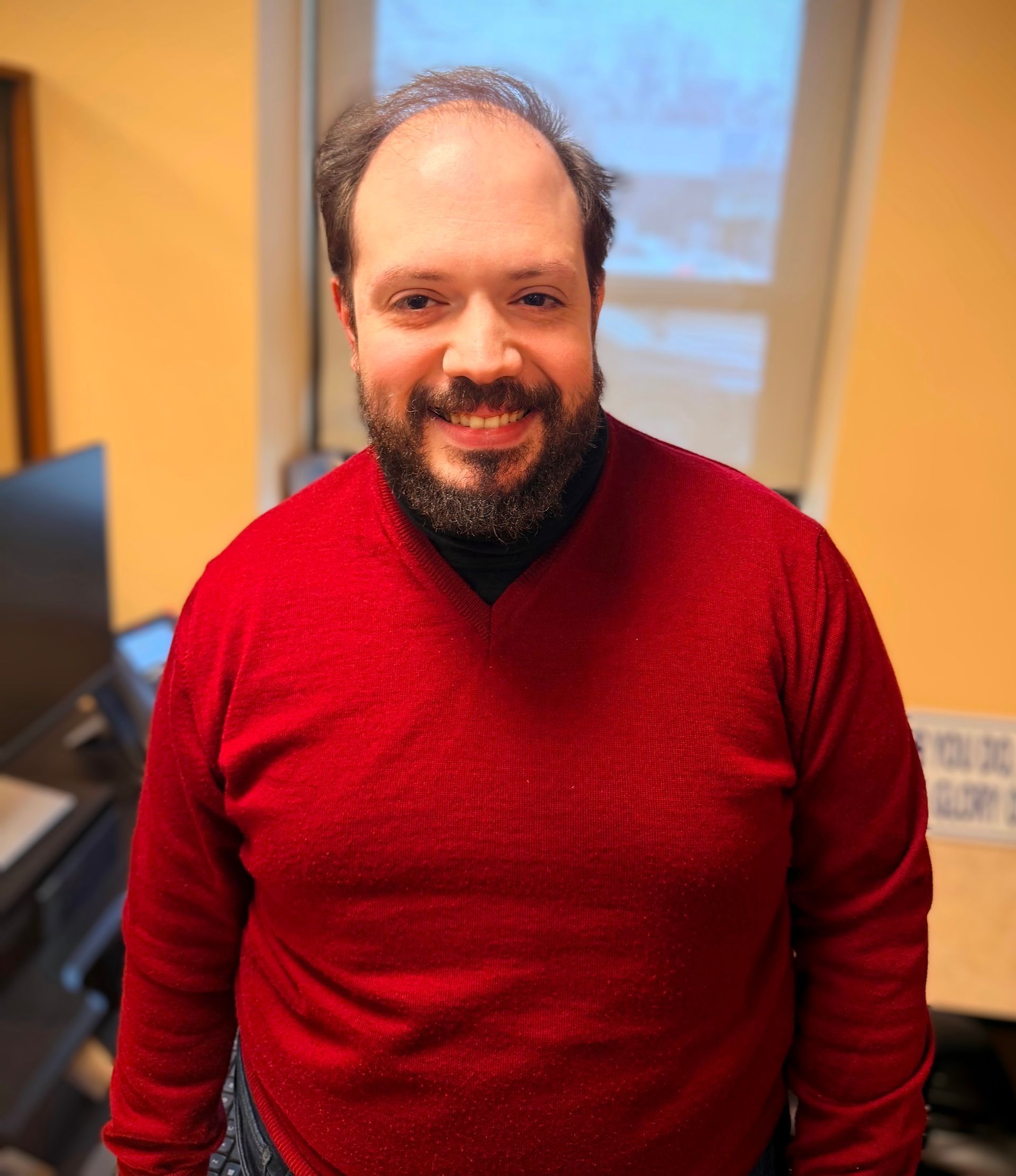 Smiling person in a red sweater standing indoors near a window.