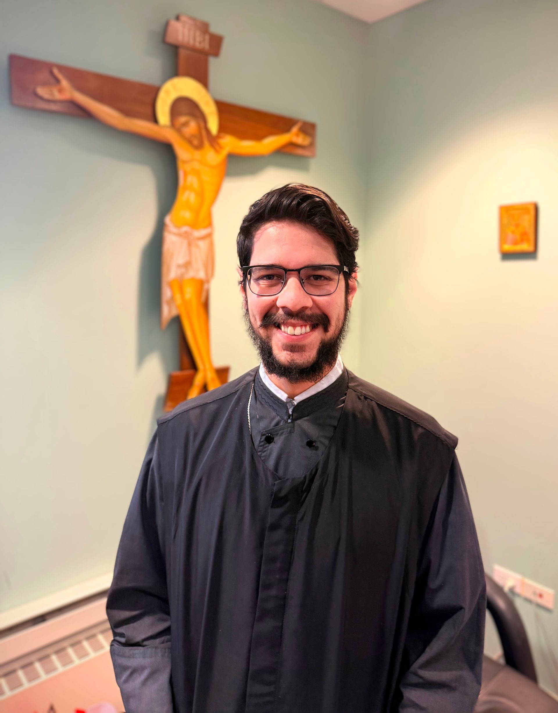 Man in black clerical shirt standing in a room with a crucifix on the wall behind him.