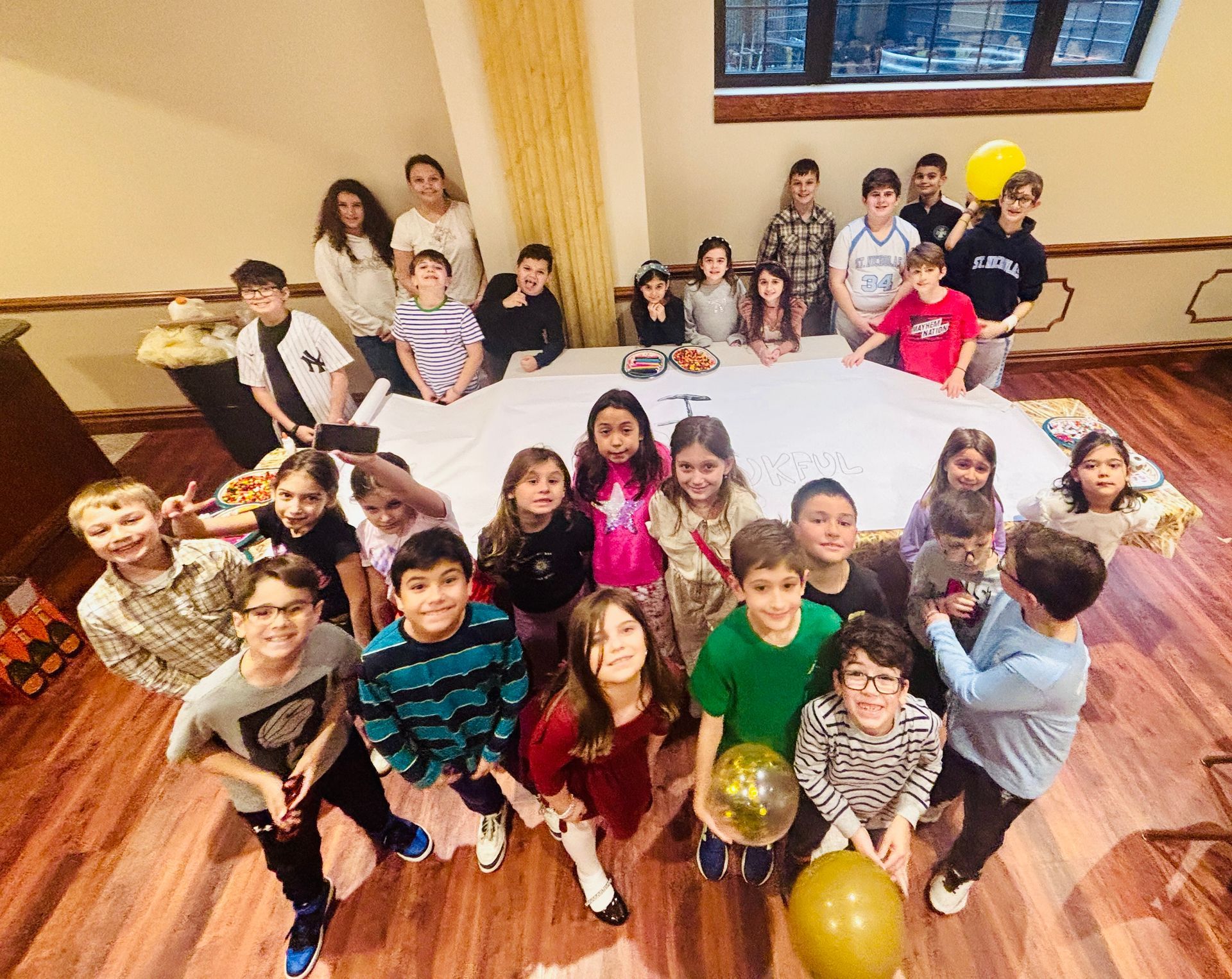 Group of children posing together indoors on a wooden floor with balloons and a few smiling toward the camera.