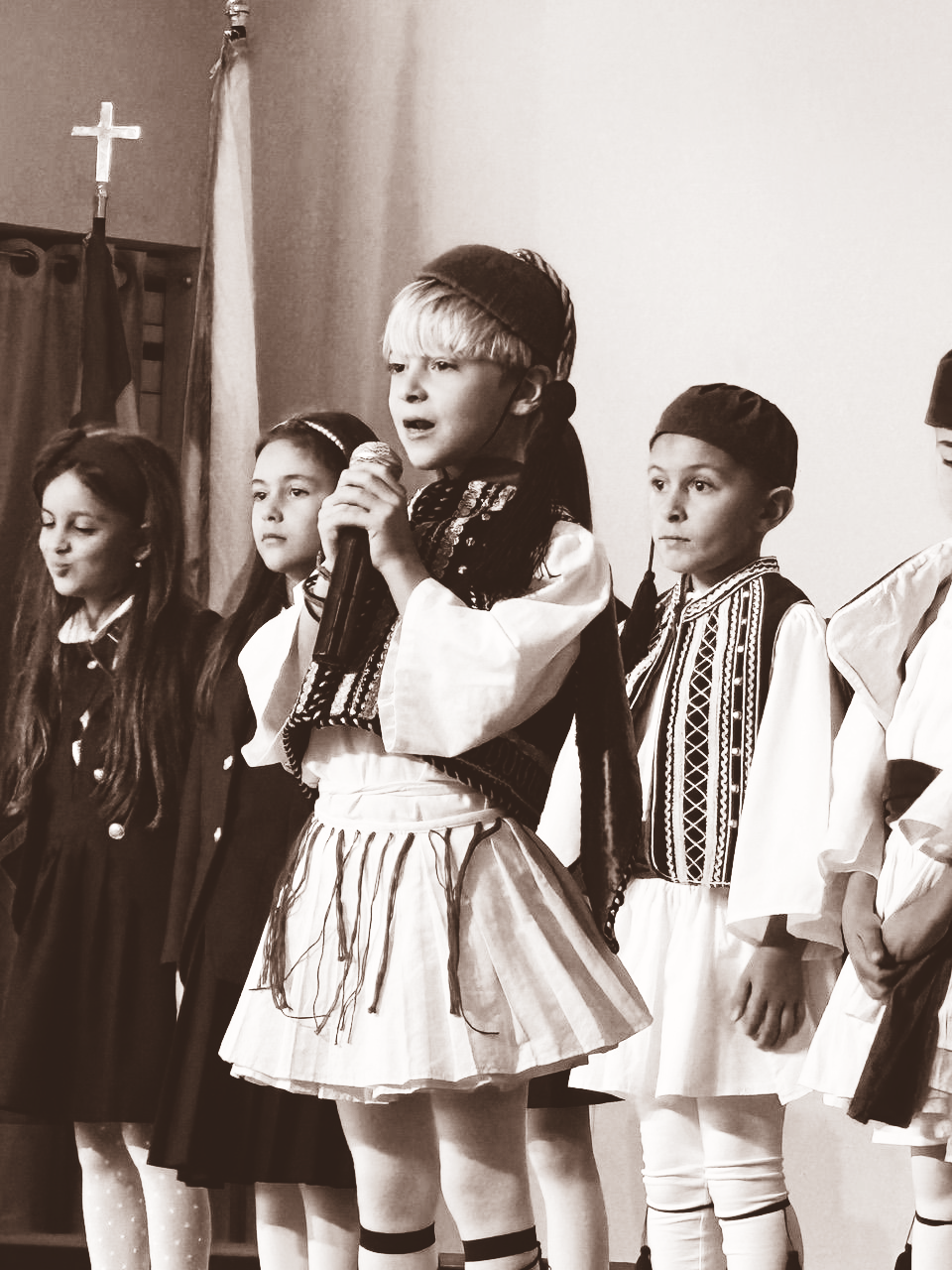 Children performing on stage, with one singing into a microphone and others standing beside her in traditional outfits