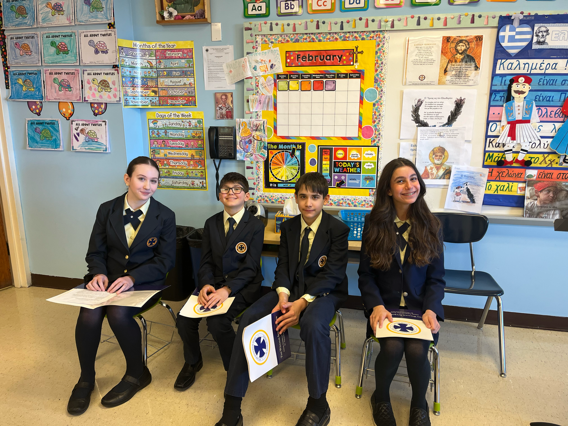 Four students in school uniforms seated in a classroom, holding papers and smiling at the camera.