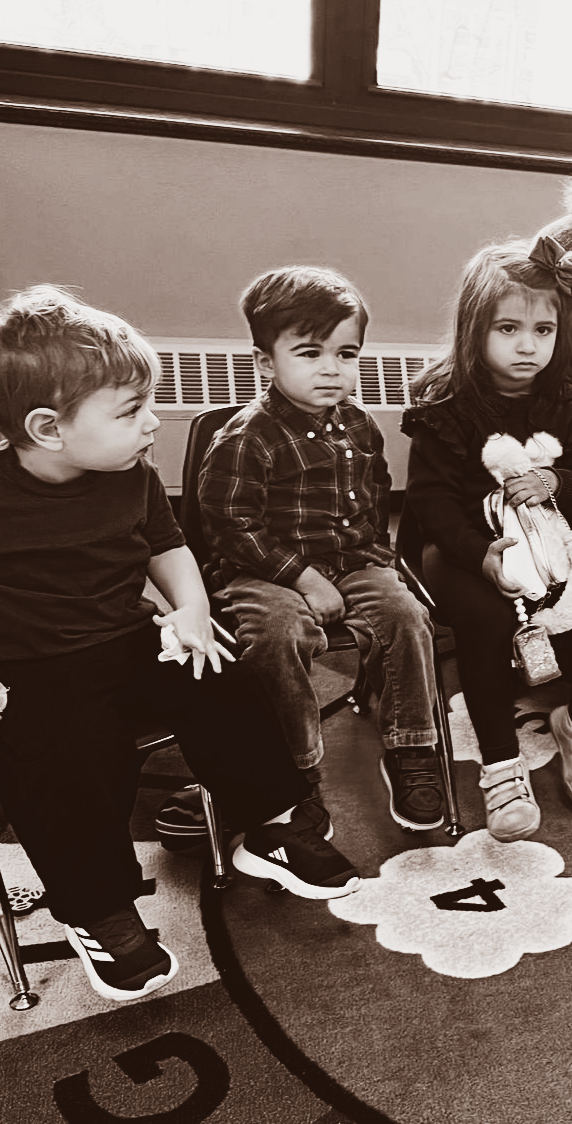 Three children seated on the floor indoors, one holding a doll, with soft window light behind them