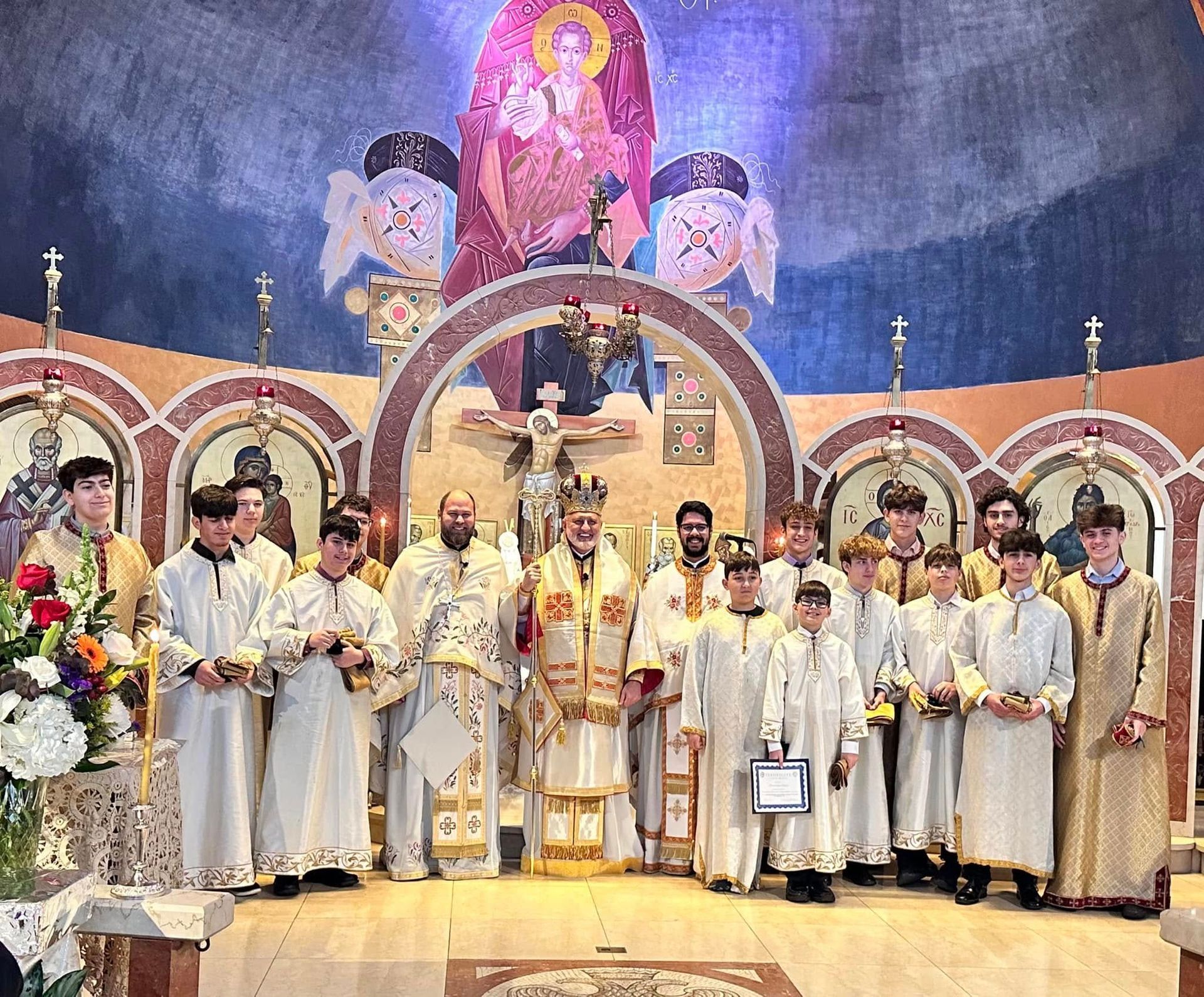 Clergy in white vestments stand on a church altar beneath ornate icons and a large mural backdrop.