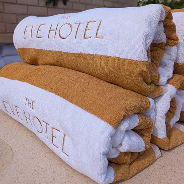 Stacked white and tan towels embroidered “EVE HOTEL” on a beige surface