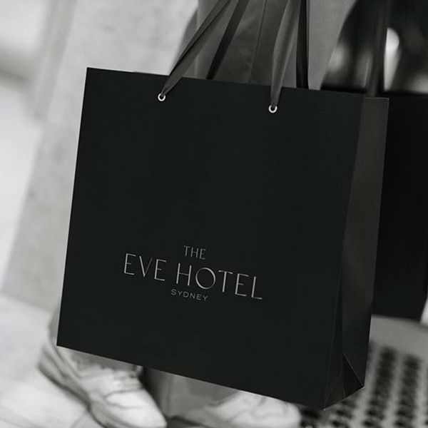 Black shopping bag labeled “The Eve Hotel Sydney” held by a person in white sneakers.