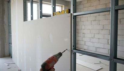 Worker installing white wall panels in a room under construction, with drill and metal framing visible