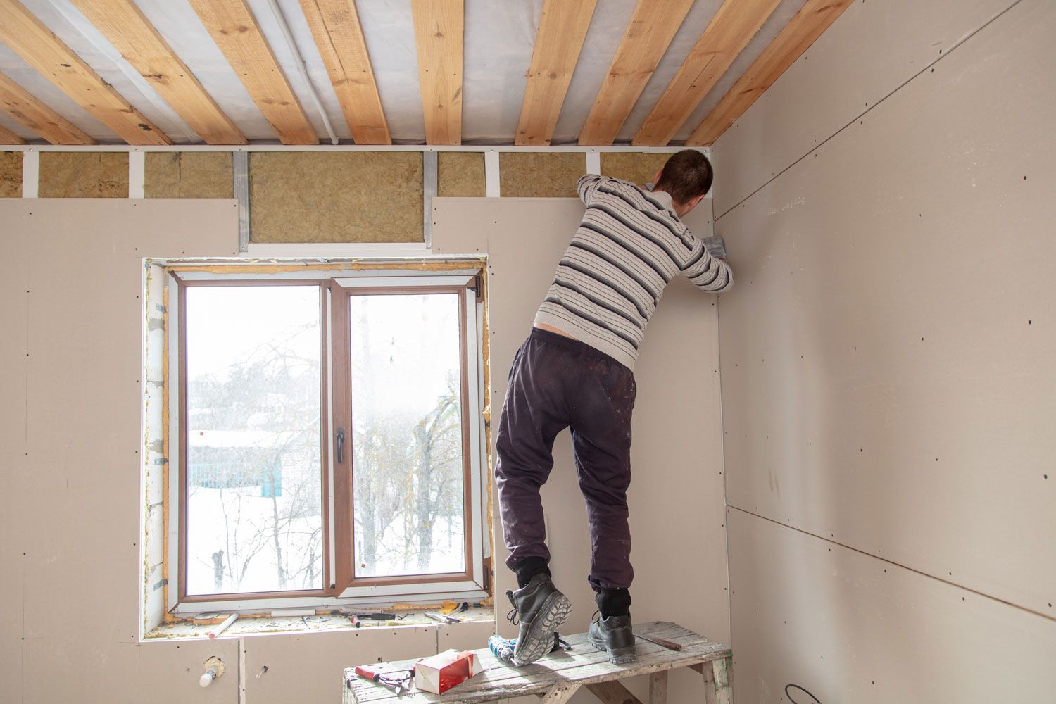 Worker installing drywall beside a window in an unfinished room Worker installing drywall beside a window in an unfinished room