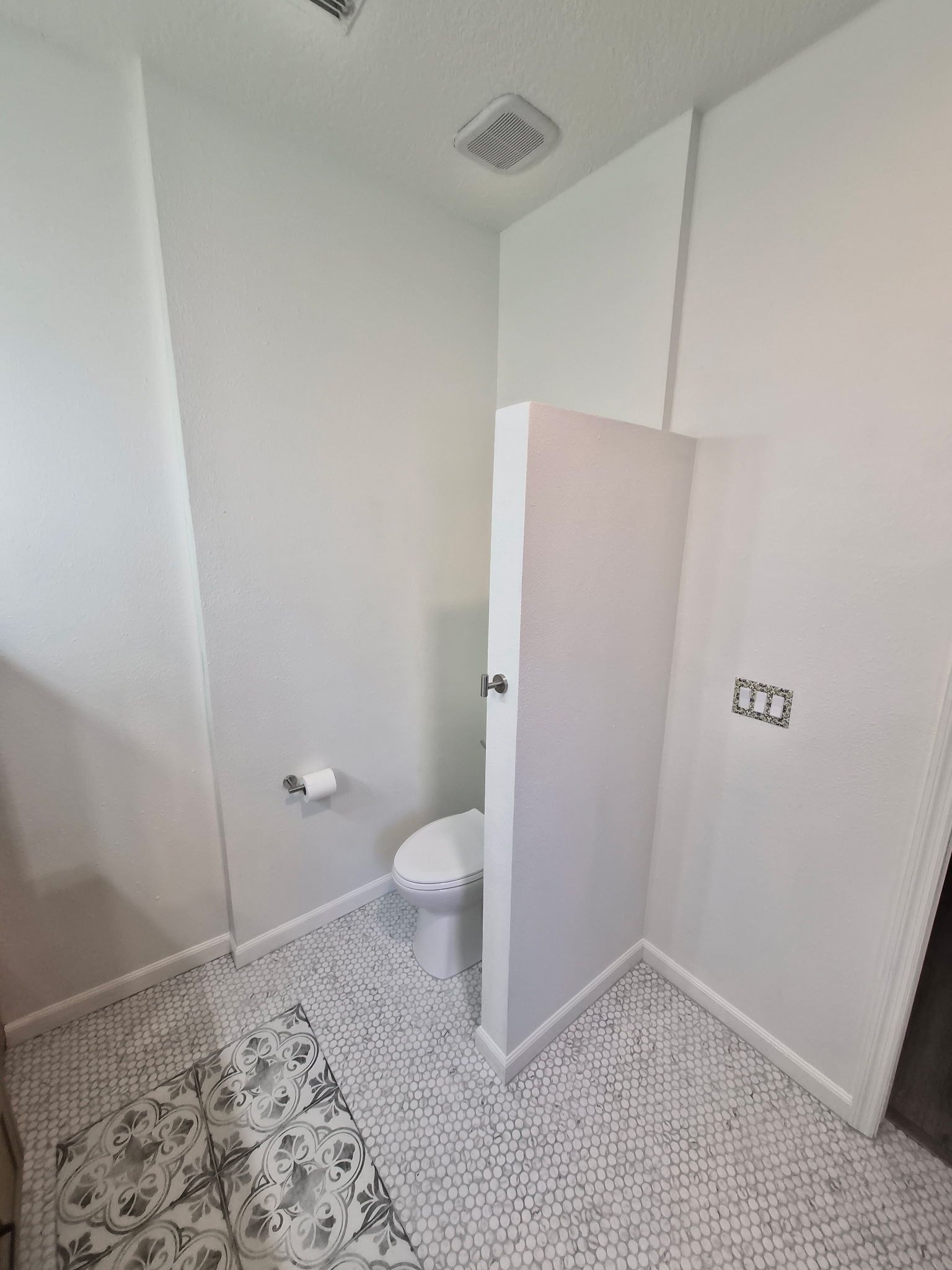 Small tiled bathroom with an open door, toilet, patterned floor tiles, and white walls.
