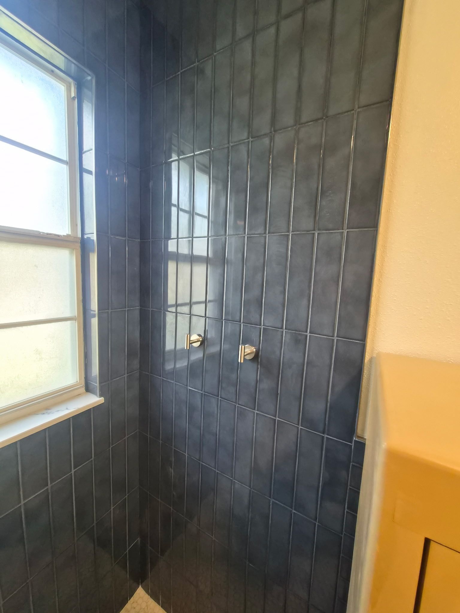 Tiled shower corner with dark wall tiles, two gold fixtures, and a window beside the shower.