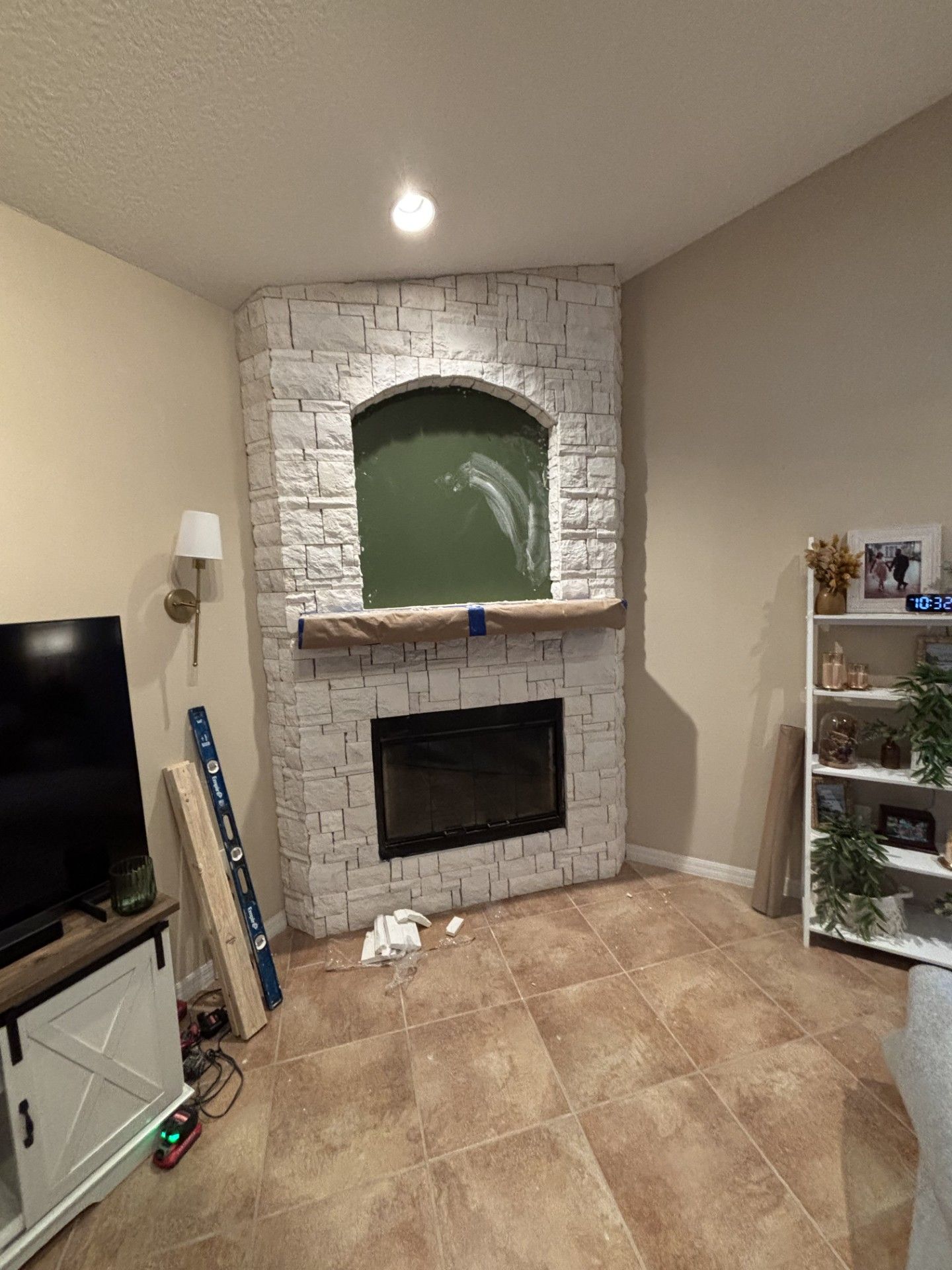 Small living room with white brick fireplace, TV on left, plant shelf on right, and tan tiled floor.