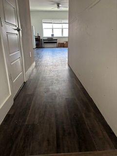 Narrow hallway with dark wood floor leading to a bright room with a table and large window.