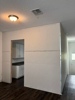 Empty room with dark wood floors, white walls, a small kitchen opening, and a hallway doorway