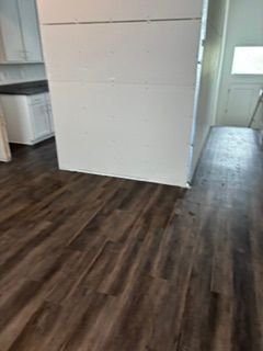 Empty room with dark wood floors, white walls, and a small kitchen area to the left.