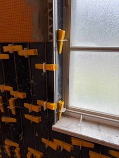 Bathroom wall with exposed tile, yellow clips, and a window ledge under renovation