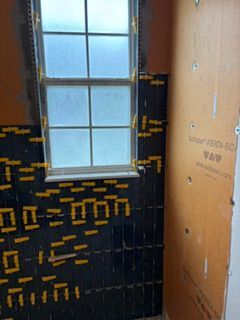 Bathroom wall under construction with black waterproofing panels around a window and tan backer board on the right.