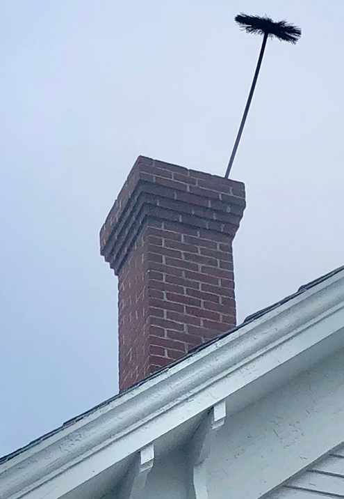 Brick chimney on a white house roof, with a black chimney brush extending upward into a gray sky.