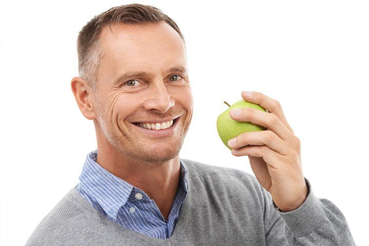 Man with gray sweater and blue shirt holding a green apple, smiling.