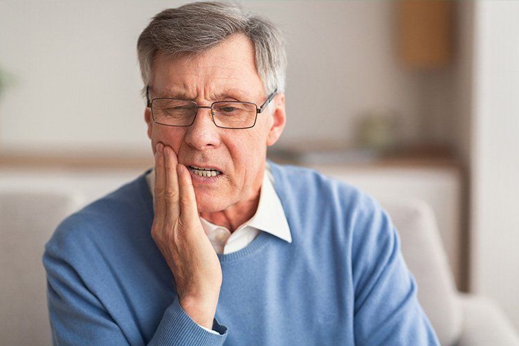 A person in a blue sweater holding their hand to their cheek with a pained expression, indicating a toothache.