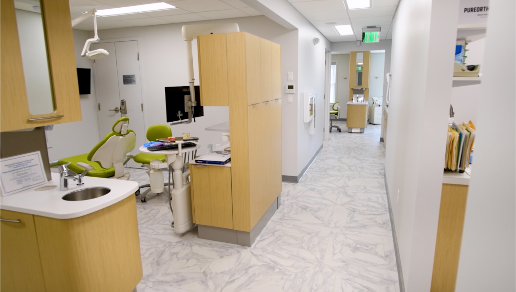 Dental office interior with a treatment room and hallway. Light wood cabinets, green dental chair, and marble-look flooring.