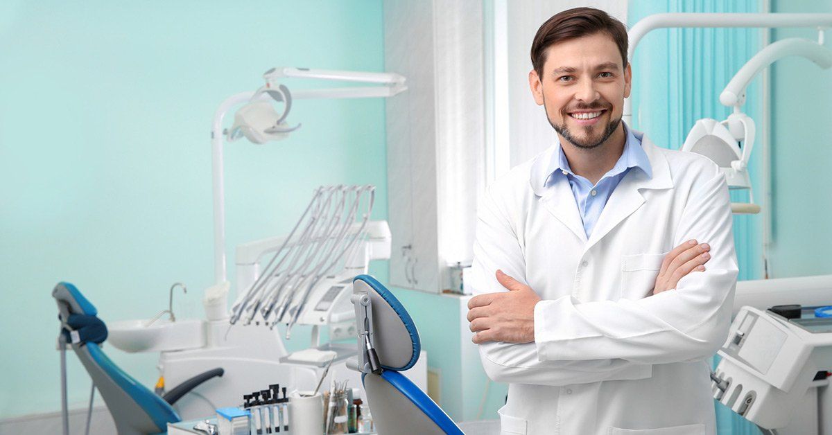A smiling dentist in a white coat stands with arms crossed in a modern, light-blue dental office.