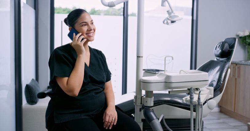 Dental assistant in black scrubs smiles while talking on a phone in a dental office.